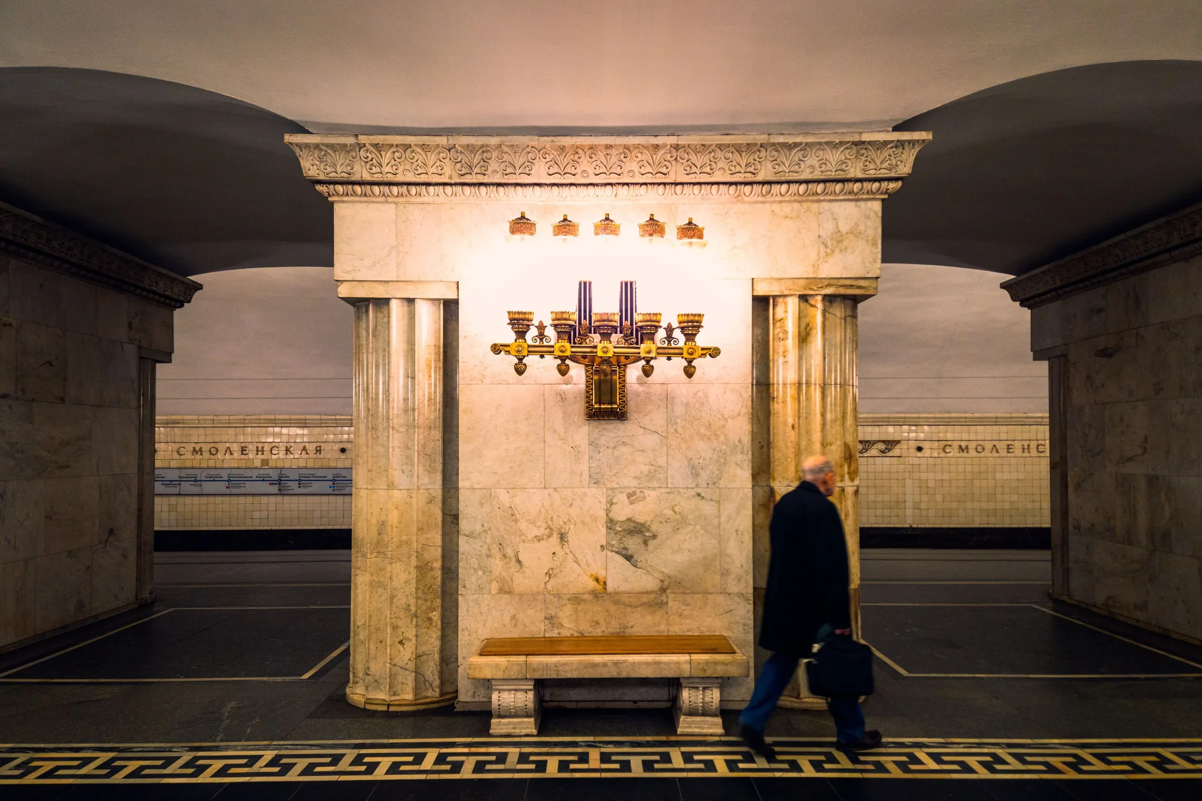 Мраморный зал московской станции метро «Смоленская», на стене бронзовый светильник, мимо проходит человек
